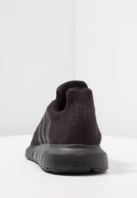 Svart stickad träningssko med texturerad sula och tre mörkare ränder på sidan, fotograferad bakifrån på vit yta.