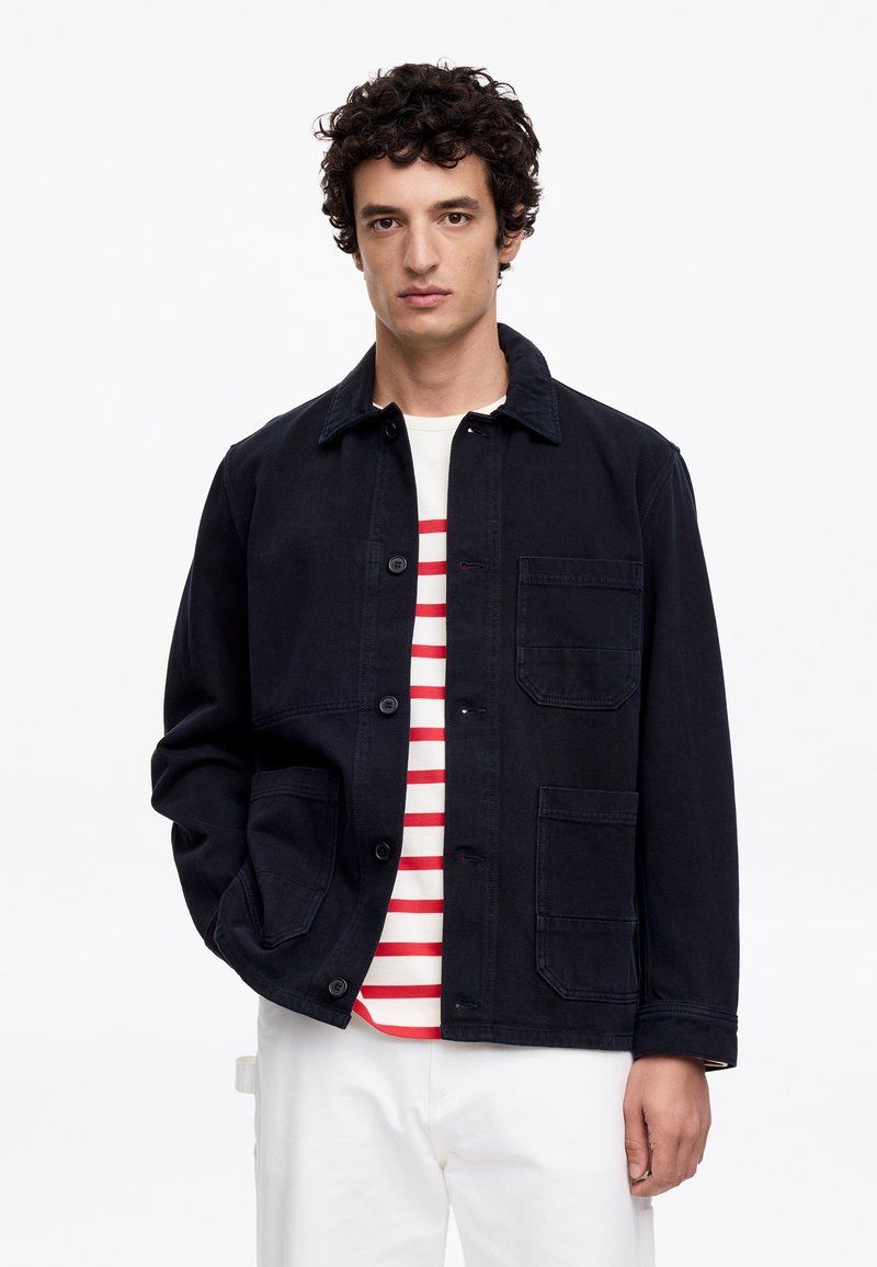 Homme aux cheveux bouclés portant une veste noire boutonnée par-dessus une chemise blanche à rayures rouges et un pantalon blanc, debout devant un fond blanc.