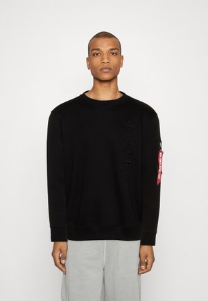 Sudadera negra con logo texturizado en la parte delantera, cuello redondo y puños acanalados. Presenta una etiqueta roja en la manga y pantalones grises.