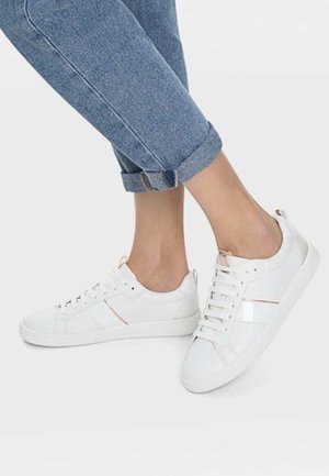 Jambes portant un jean bleu retroussé et des baskets blanches basses à lacets sur un fond clair uni.