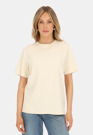 Blondynka o falowanych włosach, ubrana w beżowy t-shirt z krótkim rękawem i niebieskie dżinsy, stojąca na jednolitym białym tle.