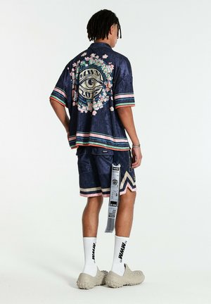 Hombre que lleva camisa azul marino y pantalones cortos con rayas rosas y verdes, diseño gráfico de ojo en la espalda, calcetines blancos y zapatos beige texturizados.