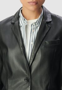 Primo piano di una persona che indossa un blazer in pelle nera sopra una camicia a maniche lunghe a righe bianche e blu e una maglietta bianca sotto.