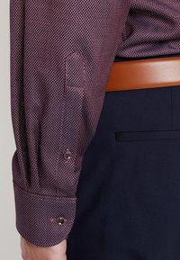 Camicia maroon a maniche lunghe con un piccolo motivo a pois, con polsini a bottoni e dettagli di cuciture rosse, abbinata a pantaloni blu scuro.
