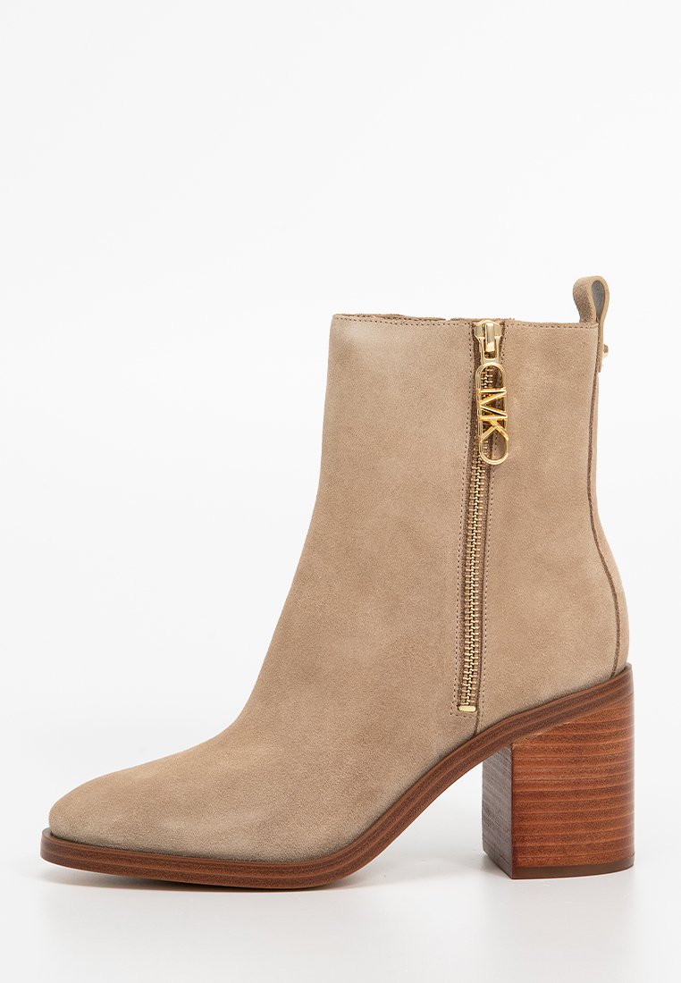 Bottine en suède beige, avec un bout pointu, une fermeture éclair sur le côté et un talon empilé marron. La fermeture éclair présente un accent doré.