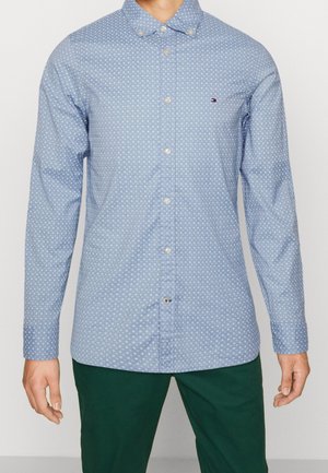 Mężczyzna w jasnoniebieskiej koszuli w białe grochy i ciemnozielonych spodniach, stojący z rozluźnionymi ramionami.