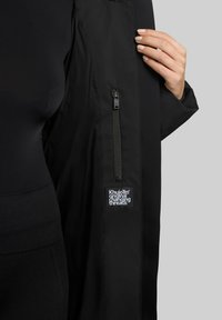 Abrigo negro con una textura suave, bolsillo lateral con cremallera y una etiqueta con "Khujo™ original changing threads" destacada.