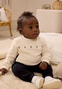 Krēmsvārtruna ar melnu "BAKER" uzrakstu, apvienota ar tumšiem biksēm un baltām kurpēm. Mīksta tekstūra uz gaiša paklāja.
