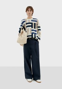 Gebreide trui met geometrische patronen in beige, marineblauw en lichtblauw, gecombineerd met brede marineblauwe broeken en een beige trekkoordtas. Witte schoenen.