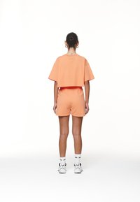Kuttet oransje t-skjorte og matchende shorts laget av myk bomull. Løs passform, enkelt design og minimale sømdetaljer. Hvite sokker og joggesko.