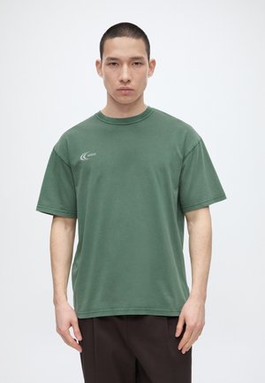 Άνδρας με κοντά μαλλιά που φοράει πράσινο T-shirt με το λογότυπο ARKK και σκούρο παντελόνι, στέκεται μπροστά σε μια απλή ανοιχτή φόντο.