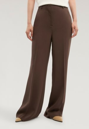 Person trägt hoch taillierte, weit geschnittene braune Hose und beigefarbene spitz zulaufende Schuhe, steht vor einem hellgrauen Hintergrund.