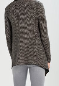 Strickjacke im braun-weißen Fischgrätenmuster, mit langen Ärmeln und einem hohen tiefen Saum, kombiniert mit grauen, eng anliegenden Jeans.