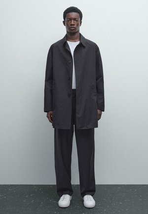 Man wearing black buttoned coat, black wide-leg pants, white shirt, and white sneakers, standing against a plain gray wall.