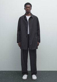 Black button-up coat with a straight cut, slightly textured fabric, front pockets, and a casual white T-shirt underneath. Worn with dark trousers and white sneakers.