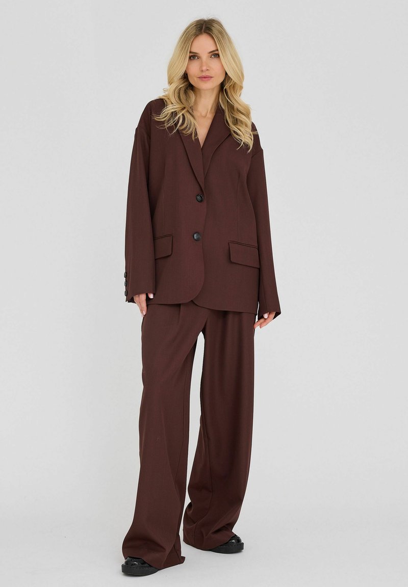 Femme blonde portant un costume marron foncé surdimensionné avec un pantalon à jambes larges et des chaussures noires, debout devant un fond clair uni.