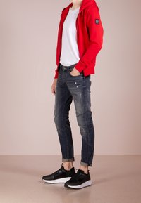 Felpa rossa con zip sopra una maglietta bianca, abbinata a jeans scuri strappati e sneaker nere con suola bianca. Design semplice e vestibilità casual.