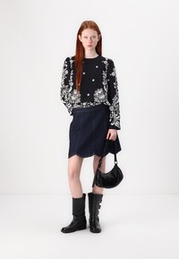 Schwarze florale Bluse mit langen Ärmeln, dunkler Denim-A-Linien-Rock mit geschwungenem Saum, schwarze kniehohe Stiefel und eine kleine schwarze Schultertasche.