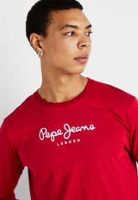 Fiatal férfi piros hosszú ujjú pólóban, rajta a "Pepe Jeans London" logóval, az oldalra néz, rövid göndör hajjal és ezüst fülbevalókkal.