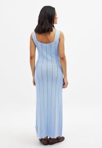 Maksidressas iš šviesiai mėlynos trikotažo medžiagos, su kvadratiniu nugara, raukšlėta tekstūra ir zigzaginiu raštu per visą ilgį. Rudos bateliai užbaigia įvaizdį.