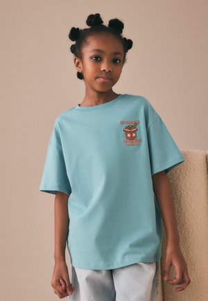 Young girl with bantu knot hairstyle wearing a light blue T-shirt with a ramen graphic and text on a beige background.