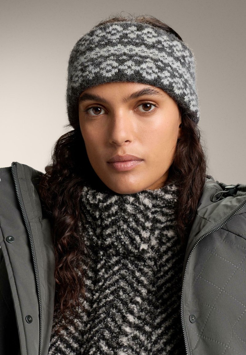 Šedý a biely vzorovaný pletený čelenka z vlnenej zmesi, s jemnou textúrou, nosená s tmavým prešívaným kabátom a vzorovaným šálom.