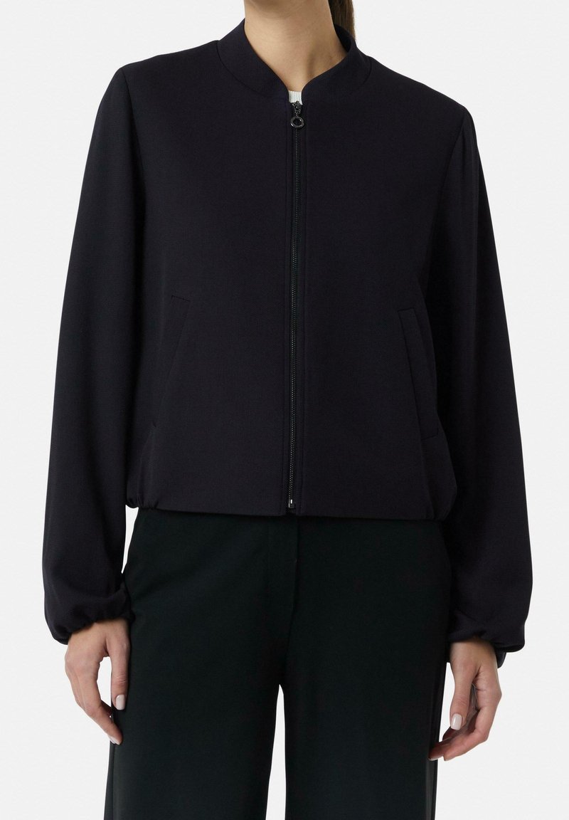 Femme portant une veste noire unie avec fermeture éclair, manches longues et poignets élastiques, associée à un pantalon foncé, debout devant un fond clair.