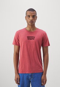 Camiseta vermelha de mangas curtas feita de tecido suave, com um gráfico do logo preto da Levi's no peito. Usada com jeans de ganga azuis.