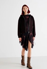 Svart zip-up hettegenser lagd over en tettsittende svart kjole med et grafisk mønster. Kombinert med brune ankelsko med spenner.