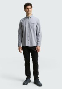 Chemise à manches longues, boutonnée, en bleu clair avec un motif à carreaux, dotée d'une poche sur la poitrine, associée à un pantalon noir et des chaussures foncées.