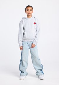 Grijze hoodie met een hartafbeelding op de borst, gecombineerd met lichtblauwe jeans met wijde pijpen en strepen aan de zijkant, en witte sneakers.