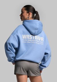 Lysblå oversized hoodie med hvid tekst på ryggen, parret med grå shorts. Blødt stof, snørelukning i hætten og afslappede ærmer.