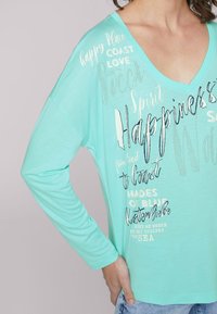 Myntgrön långärmad skjorta med V-ringning. Har tryckt text i olika teckensnitt, varav några har metallicdetaljer. Mjuk, slät textur.