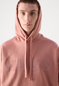 Rosa Kapuzenpullover mit einem großen, geprägt "HUGO" Logo. Verfügt über eine Kordelzugkapuze und eine weiche Stofftextur. Sauberes, minimales Design.