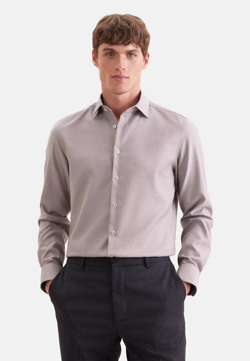 Jeune homme portant une chemise gris clair à manches longues boutonnée et un pantalon foncé, les mains dans les poches, face à l'avant.