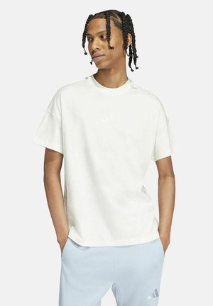 T-shirt bianca in cotone con maniche corte, scollo rotondo, vestibilità rilassata, caratterizzata da un sottile logo Adidas sul petto. Abbinata a pantaloni azzurri chiari.
