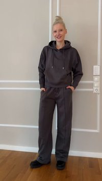 Personne souriante aux cheveux blonds attachés en chignon, portant un sweat à capuche gris foncé et un pantalon large assorti, debout devant un mur beige avec une moulure blanche.