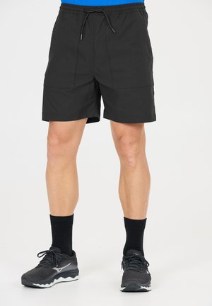 Mann trägt schwarze Sportshorts, schwarze Sportsocken und schwarze Laufschuhe, steht vor weißem Hintergrund.