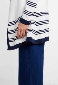 Biało-granatowy sweter w paski z dzianiny z okrągłym dekoltem i długimi rękawami, nałożony na ciemnogranatowe dżinsy w szerokim kroju.