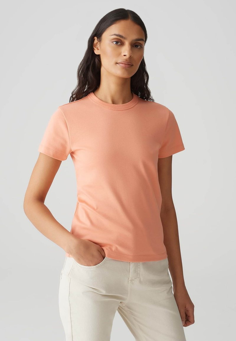 T-shirt a maniche corte con collo rotondo in morbido tessuto pesca, dal design aderente e texture liscia, abbinato a pantaloni chiari.