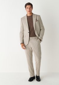 Costume gris clair en tissu texturé, associé à un pull marron. Le costume présente une fermeture à un seul bouton et des chaussures noires.