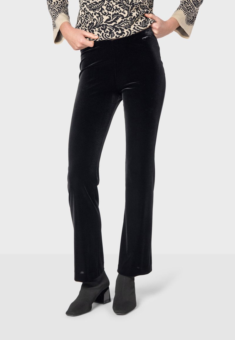 Čierne zamatové nohavice so štíhlym strihom a jemným rozšírením na spodnej časti, s hladkou textúrou a vysokým pásom. V kombinácii s členkovými topánkami.