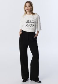 Camicetta bianca con texture e la scritta "MERCI AMOUR" stampata in nero, abbinata a pantaloni neri a gamba larga, con tasche laterali e taglio sartoriale.