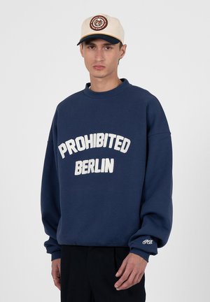 Giovane uomo che indossa un cappellino beige e una felpa blu navy con la scritta "PROHIBITED BERLIN" sul davanti e iniziali ricamate sulla manica.