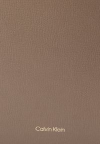 Barna textúrázott bőrfelület, az alján jobbra aranyszínű "Calvin Klein" logóval.