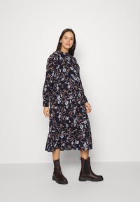 Vestido de manga longa com estampa floral num fundo azul marinho escuro, com bainha ondulada, combinado com botas de tornozelo castanhas com sola grossa.