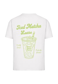Balta marškinėliai su žaliu piešiniu, vaizduojančiu šaltą matcha latte su šiaudeliu, apsuptu tekstų: "Šaltas matcha latte", "Šviežiai malti" ir "Puikiai išplakti".