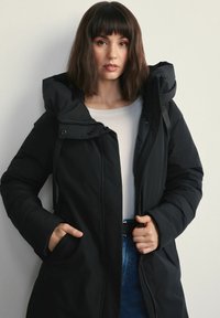 Kobieta z brązowymi włosami sięgającymi ramion, ubrana w czarną kurtkę puchową na białą bluzkę i niebieskie dżinsy, stojąca na tle jasnego, gładkiego tła.