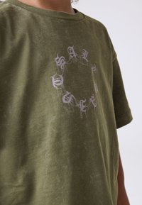 Person som bär en olivgrön t-shirt med en bleknad lila cirkulär text i gotisk stil på bröstet mot en enfärgad bakgrund.