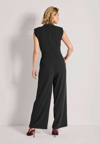 Zwarte jumpsuit met kapmouwen, getailleerd lijfje en wijde pijpen. Voorzien van een ceintuurtaille en gladde stof, gecombineerd met rode hoge hakken.